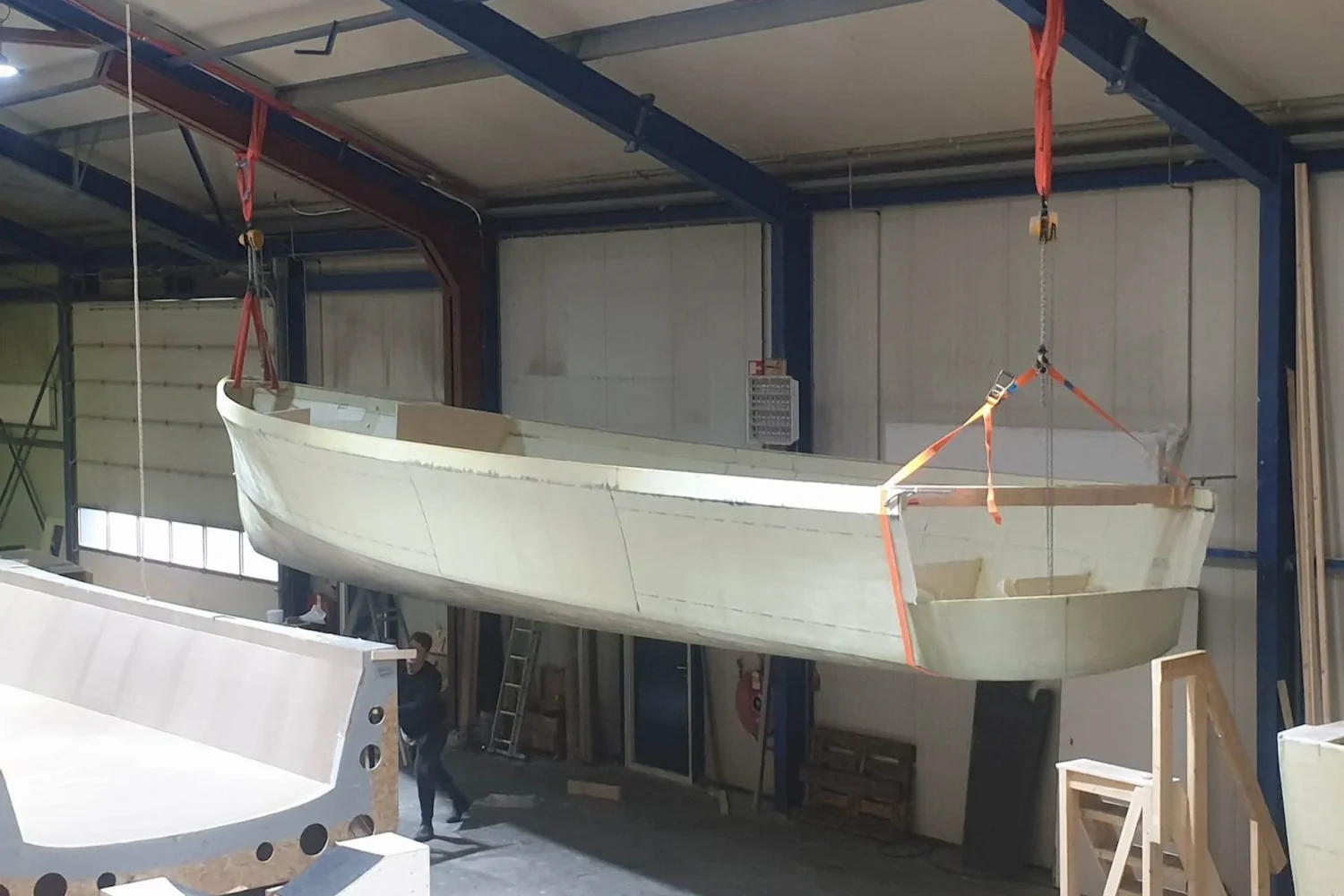 Light-coloured boat hull suspended from a crane in the workshop